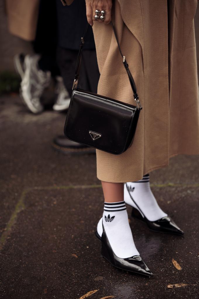 A guest at Paris Fashion Week, wearing white Adidas half-crew socks, black Prada shoes, and holding a black Prada bag