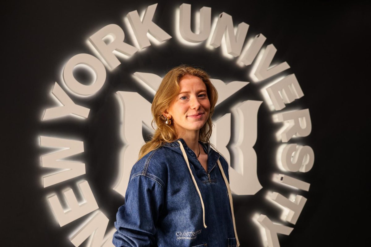 A girl wears a blue jean sweater and poses for a portrait in front of a white illuminated sign that reads “New York University” in a circular shape with the N.Y.U. Athletics logo in the center.