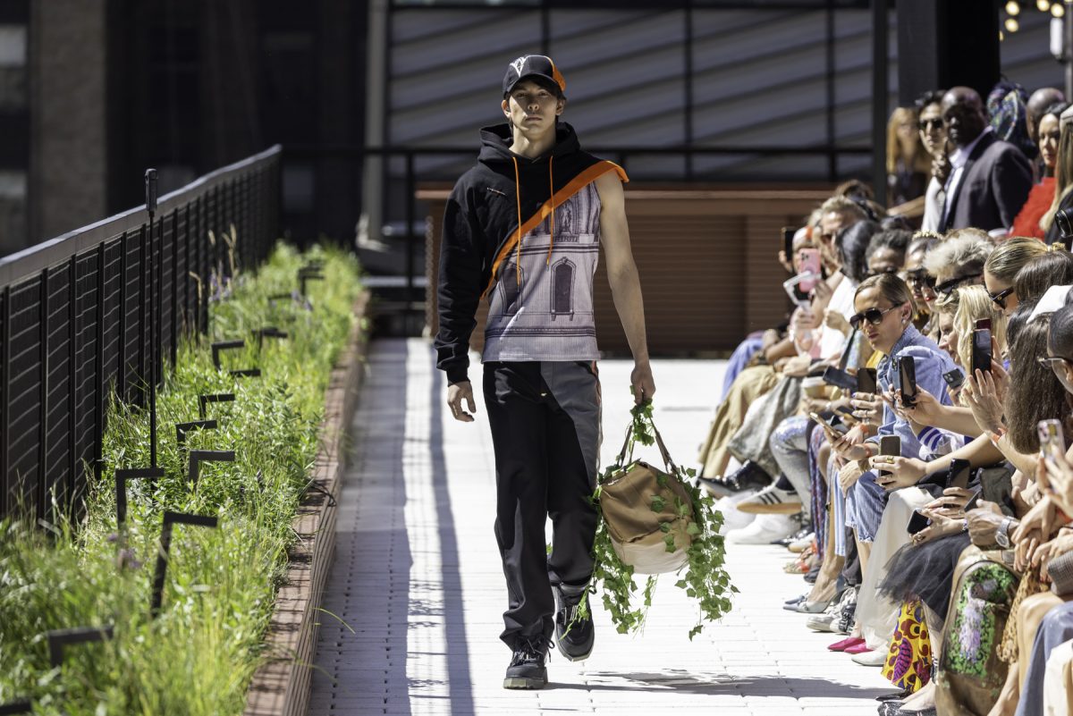 A model wearing a black hat and hoodie with orange accents, a white undershirt and black pants and shoes walks down a white tiled runway while carrying a beige bag with green leaves.