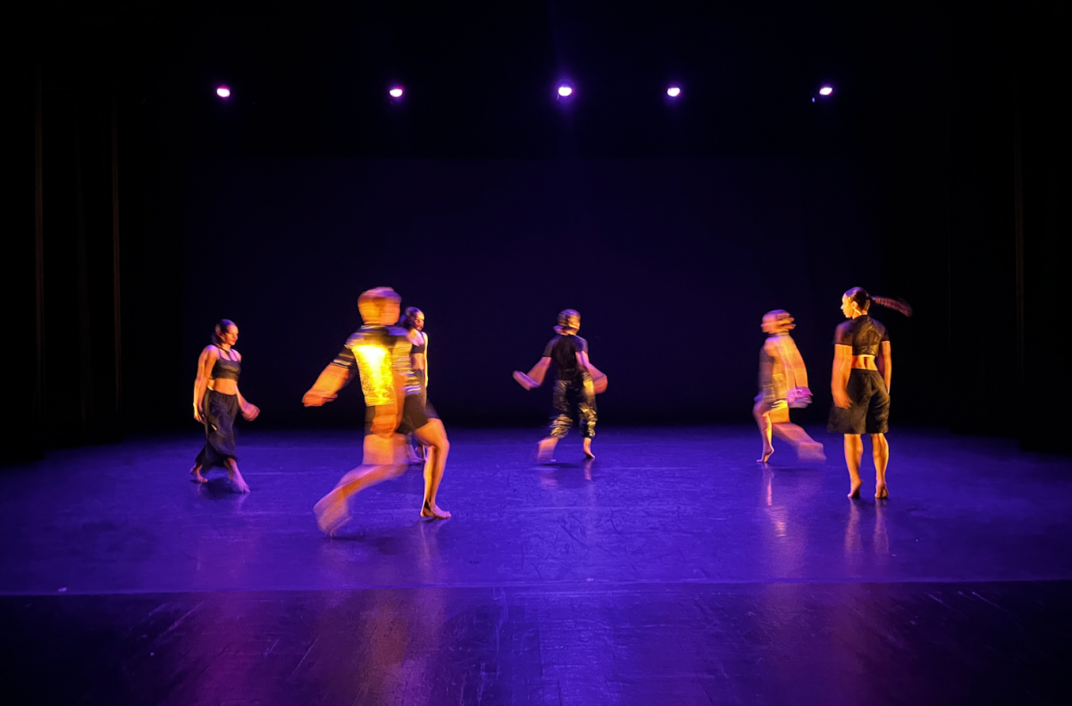 Group of dancers performing on a stage with purple lighting.