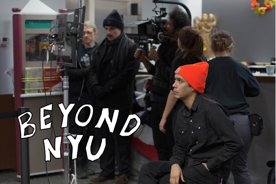 A man wearing an all black outfit and a bright orange beanie watches a monitor. Behind him are crew members wearing black, one of whom is holding a camera. 