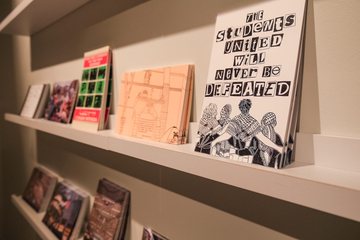 Several postcards on a shelf. The closest one reads “THE STUDENTS UNITED WILL NEVER BE DEFEATED” in block text, with a black and white drawing of people wearing keffiyehs with their arms around each other.