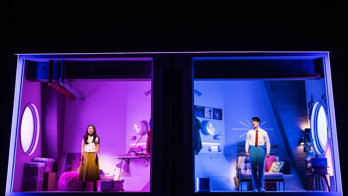 A man and woman stand facing the audience in adjacent rooms on a stage. The woman’s has a purple hue while the man’s is blue.