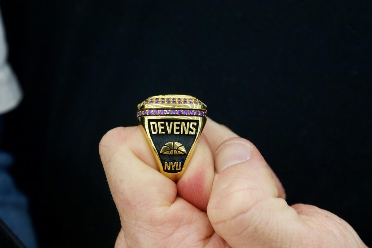 A hand is folded to show off a gold and black ring with purple gems. The ring says “DEVENS” and “N.Y.U.” and has the silhouette of half of a basketball.