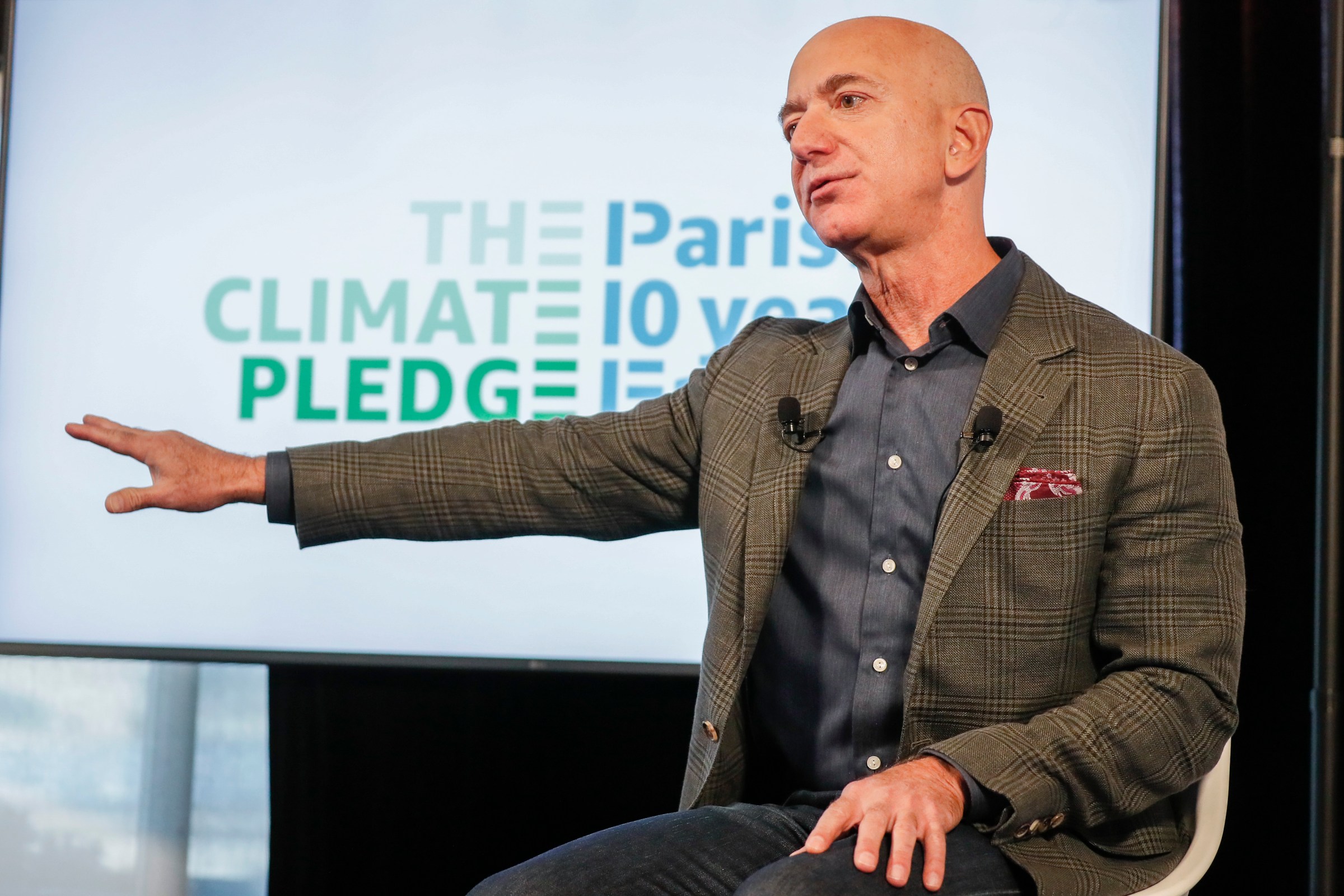 Amazon CEO Jeff Bezos sitting down onstage and speaking with his right arm extended at his side in front of a “Climate Pledge” logo.