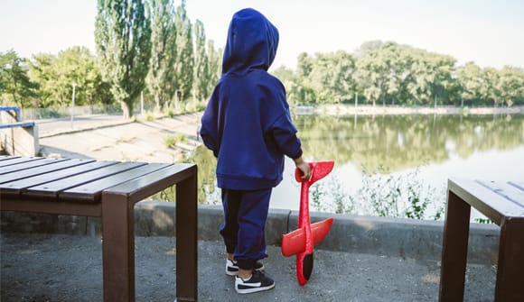 photo of the back of a child wearing a blue hoodie and holding a red toy airplane looking out over a pond