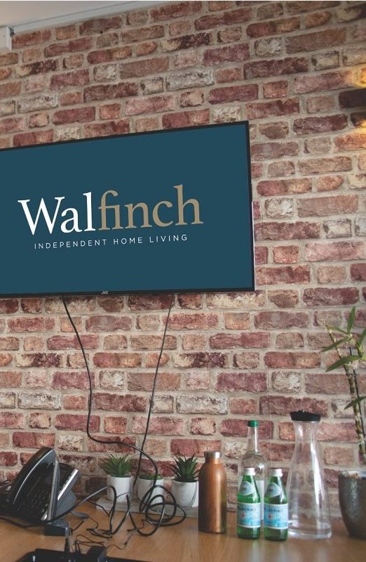 Internal brick wall with desk in front at the bottom. A tv screen is hung on the wall showing the green Walfinch logo. There are waterbottles and a landline phone on the desk.