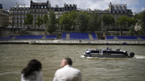 Des personnes sont assises le long de la Seine, face à des tribunes installées sur ses berges pour la cérémonie d'ouverture des Jeux olympiques de Paris, le 4 juillet 2024.