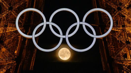 لقطة تظهر القمر أثناء تداخل انعكاسه مع الحلقات الأولمبية المعروضة على برج إيفل في 23 تموز/يوليو 2024 في باريس، قبل أيام قليلة من حفل افتتاح دورة الألعاب الأولمبية 2024 في باريس.