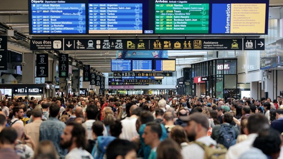 Passageiros reunidos em redor dos placards de partida e chegada da estação Montparnasse em Paris, a 26 de Julho de 2024, numa altura em que a rede ferroviária TGV de França é atingida por actos de sabotagem que perturbaram o sistema, escassas horas antes da cerimónia de abertura dos Jogos de Paris 2024.