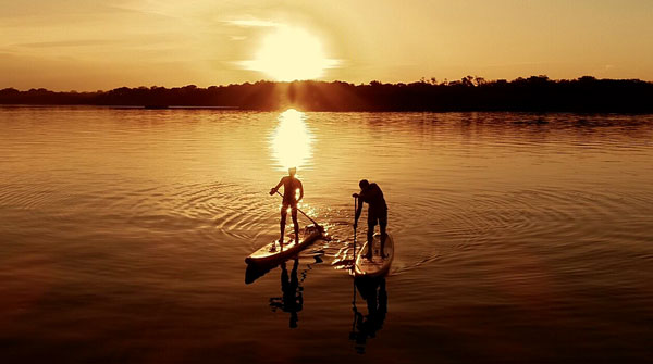 Dois praticantes de Stand Up Paddle em um rio ao por do sol