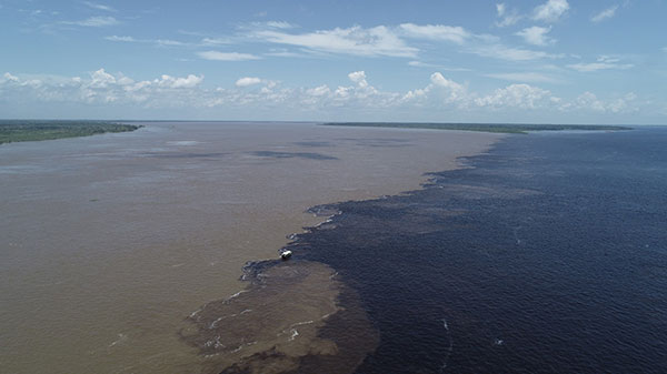 Foto de um extenso rio, com águas metade claras e metade escuras, com um pequeno barco ao meio