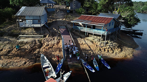 Foto aérea do cais de um povoado, com dois barco estacionado e quatro pranchas de standup. Na beira do cais há parte da população do vilarejo.