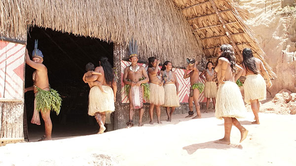 Foto da entrada da oca, com índios ao redor. Um índio escora-se na porta à esquerda, uma índia entra na oca segurando um bebê. Cinco jovens, 2 índios e 3 índias, estão em pé na parede externa da oca conversando. Duas índias jovens estão caminhando em direção a oca.