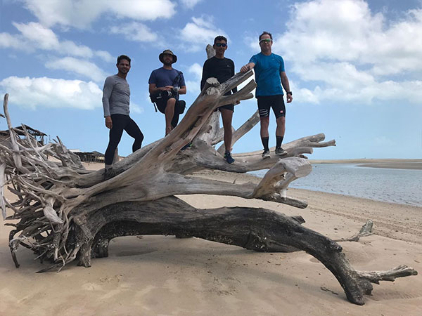 Conservai e mais três homens possam em cima de uma árvore caída na praia.