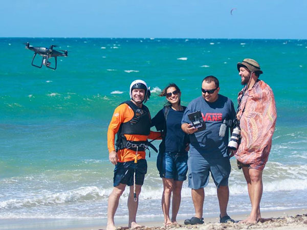 Apresentador Conservani junto com três pessoas na praia ao lado de um drone.