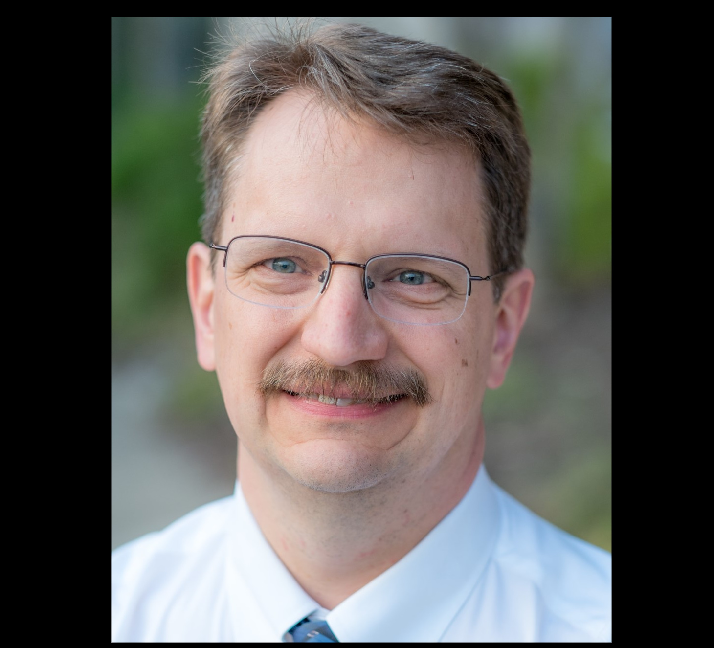 Jonathan Gardner smiles at the camera in a professional portrait. He wears glasses, a white shirt and blue tie. The background is blurred and green.