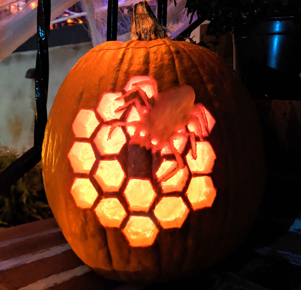 View of an illuminated jack-o'-lantern style pumpkin carved with an image of the 18 hexagon mirror segments of the James Webb Space Telescope with a spider figure in the top right portion of the overall collection of mirror segments.