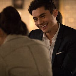 In a scene from “Crazy Rich Asians,” Henry Golding, wearing a navy suit and a white shirt with the top button undone, sits at a bar talking to Constance Wu, who faces him.