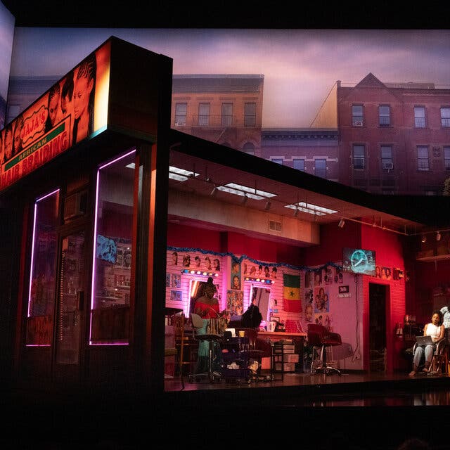 A scene from the play “Jaja’s African Hair Braiding” shows a rectangular set underneath a projection of city buildings.