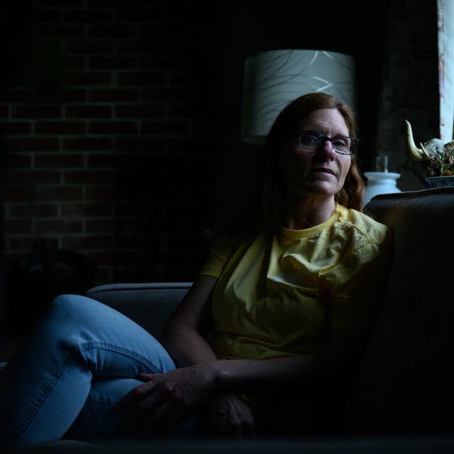 Kim Jones sits in her living room covered in shadows. She wears a yellow t-shirt and jeans.