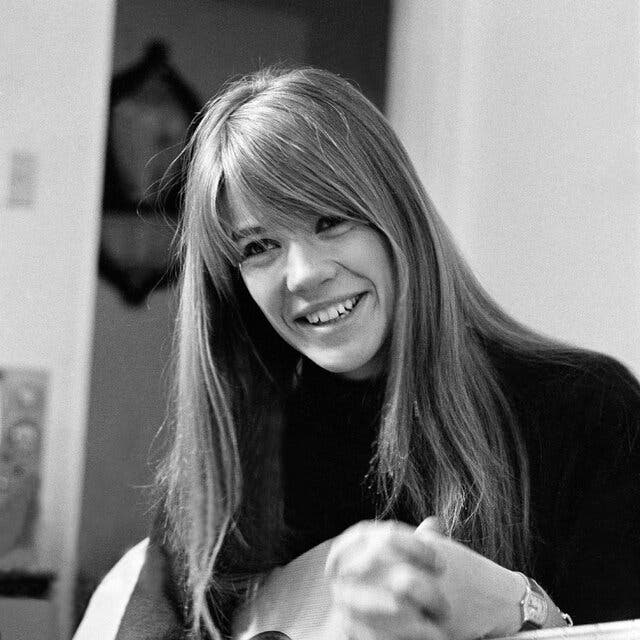 Françoise Hardy holds a guitar and smiles, her long hair falling over her shoulders.