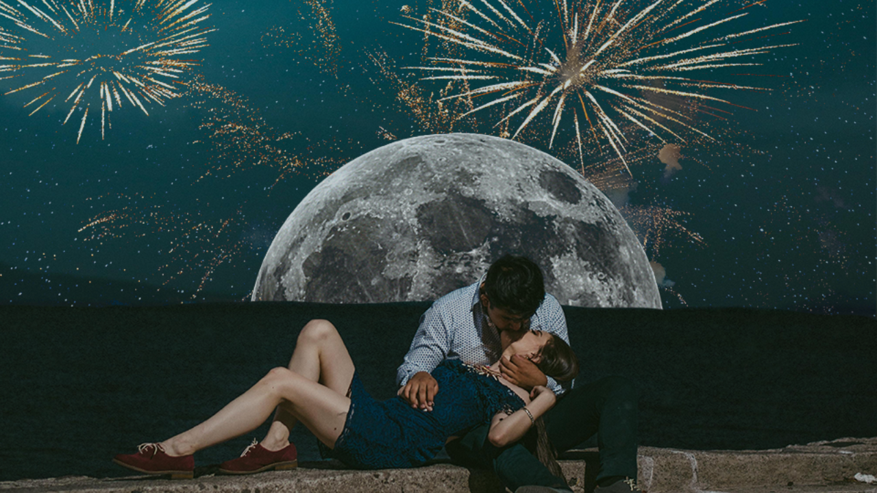 A man and a woman sharing a kiss by the beach, beneath a night sky filled with fireworks and a full moon