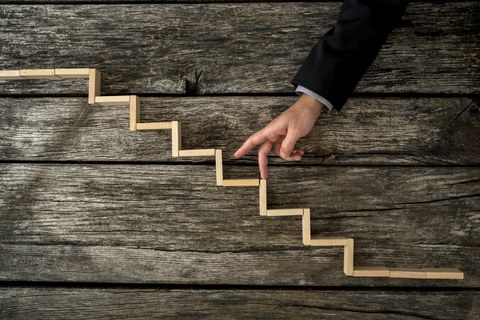 Das Foto zeigt einen Arm vor einem hölzernen Hintergrund. Darauf liegen Holzstückchen, die eine Treppe bilden. Die Person geht mit ihren Fingern die Treppe hinauf.