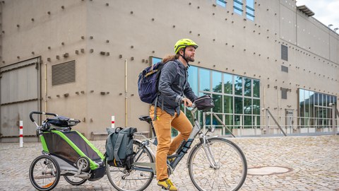 Foto eines jungen Vaters auf einem Fahrrad, der mit einem Kind hinten im Fahrradanhänger über den TU Campus fährt. 