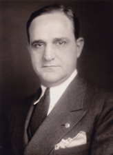 A half-length portrait of a man wearing a suit with a smirk on his face and a slight purple hue about him