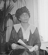 Woman in a dark hat holding a book sits in front of a fronded plant. She is in a white collared shirt and her head is slightly cocked