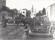 City buildings, some with people looking down from roofs, people and soldiers in a large square, with motor car in foreground