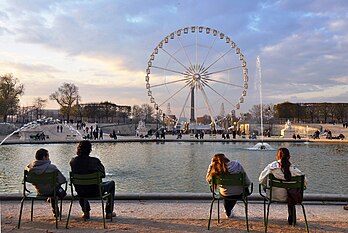 Pessoas sentadas nos jardins das Tulherias, em frente à Roda de Paris, França. Essa roda-gigante transportável de 60 metros de altura foi originalmente instalada na Praça da Concórdia para as celebrações do milênio de 2000. Saiu de Paris em 2002 e desde então tem prestado serviços em vários outros locais ao redor do mundo. Devido ao seu design transportável, pode ser montada em 72 horas e desmontada em 60 horas por uma equipe especializada. As quarenta e duas cabines podem ser carregadas três ou seis de cada vez, e cada uma pode acomodar oito passageiros. (definição 4 117 × 2 755)