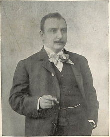 Photographie d'un homme moustachu, en costume, nœud papillon et cigare à la main droite.