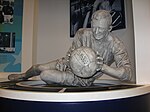 Photographie montrant une statue au musée de Manchester City représentant Bert Trautmann en équipement de gardien de but, couché, tenant un ballon de football des deux mains