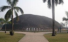 Sebuah bukit yang sangat besar di belakang dua pohon palem dan sebuah bjalan raya, pejalan kaki hanya 1/5 dari tinggi bukit