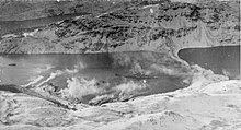 Black and white photograph of a ship in a bay. Smoke is drifting up from the land.