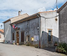 Photo de la mairie, un bâtiment de plein pied avec trois drapeaux au dessus de la porte (région Occitanie, France, Union européenne). D'un côté du bâtiment, on distingue un panneau d'affichage et une boîte aux lettres de la Poste, de l'autre côté les affiches de l'élection présidentielle de 2022 posés au niveau du sol.