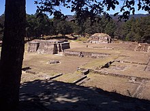 Vista de un número de ruinas bajas, bien mantenidos, que consisten en un laberinto de plataformas rectangulares basales superpuestas. Dos pequeñas estructuras piramidales dominan la vista, con un fondo de bosque de pinos.