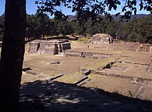 Vista d'un nombre de ruïnes baixes, ben mantingudes, que consisteixen en un laberint de plataformes rectangulars basals superposades. Dues petites estructures piramidals dominen la vista, amb un fons de bosc de pins.