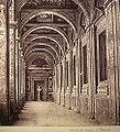Schwarzweißfotografie von einer Loggia, die auf der linken Seite offene Säulen und auf der rechten Seite eine geschlossene Außenfassade mit Fenstern hat. An der Decke sind Malereien mit Rundbögen dazwischen, die die Säulen verbinden. Am Ende des Ganges steht eine Büste auf einem Podest.