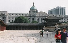 Photographie prise depuis l'intérieur d'un ancien palais coréen. L'enceinte extérieure est le principal élément visible, avec plus loin à l'extérieur un grand bâtiment japonais qui domine l'ensemble.