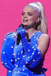 Margaret performing on-stage in a blue dress on a pink background.