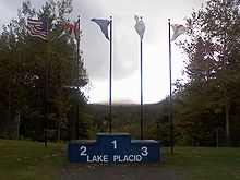 Un podium portant l'inscription « Lake Placid » devant cinq drapeaux.