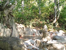 Photo couleur montrant trois hommes assis dans un bain thermal en extérieur avec une forêt en arrière-plan.