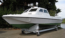 View of a small streamlined boat resting in a cradle out of the water. The boat is painted white and green, with "G 718" painted on the side. Two searchlights and a loudhailer are attached to the roof of its open-backed cabin.