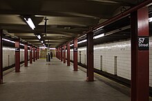 Blick entlang des Mittelbahnsteigs der Bahnhofs 57th Street. Der Raumeindruck wird bestimmt von den cremefarben gefliesten, teils schmutzigen Hintergleiswänden, den massiven dunkel-weinroten T-Trägern, die als Stützenreihen in der Nähe der beiden Bahnsteigkanten verlaufen, von den dunkel angelaufenen Betonplatten des Bahnsteigbodens und von einer dunkelbeige angestrichenen Decke aus quer verlaufenden Stahlträgern und vermutlich Betonfüllungen. Entlang der Decke an den Bahnsteigkanten verlaufen ununterbrochene Bänder von Leuchtstoffröhren, von denen aber manche defekt sind und nicht leuchten. In der Bahnsteigmitte ist eine dritte, allerdings aus Einzelröhren mit jeweils Lücken zwischen den Leuchtstofflampenhaltern bestehende Lichtreihe. Im Bildhintergrund blickt man direkt auf zwei jeweils die halbe Bahnsteigbreite einnehmende Treppen, die vom Bahnsteig nach oben führen. An jeder zweiten der dunkelroten Bahnsteigstützen sind Schilder mit dem Bahnhofsnamen montiert im typischen Transit-Authority-Design in der Schreibweise „57 Street“ mit weißer Schrift auf schwarzem Grund. An den Hintergleiswänden ist der Bahnhofsname in regelmäßigen Abständen aufgedruckt in der Schreibweise „57th Street“.