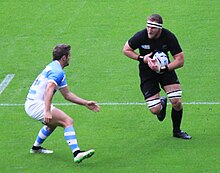 Joueur de rugby de face en noir, tenant le ballon sous son bras grauche, un adversaire sur sa droite s'appretant à le bloquer.