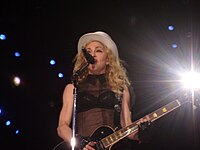 Picture of a middle-aged blond woman uptill the waiste, singing in front of a microphone. Her hair is in waves and falls up to her shoulders. She appears to be wearing a black bra covered with a sleeveless netted covering and wears a white hat on her head. There are black gloves on her hand and she plays an electric guitar. Behind her, to her left, a flood light is visible.