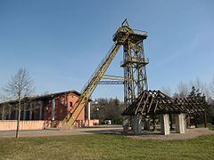 Reconstitution d'une machine à molette datant de 1790 (premier plan), l'ancien chevalement du puits Sainte-Marie (mis en place en février-mars 1988) et la mairie.